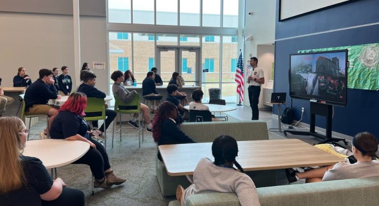 A group of students sit and watch a person giving a presentation next to an American flag and a large screen in a bright, modern classroom with large windows.