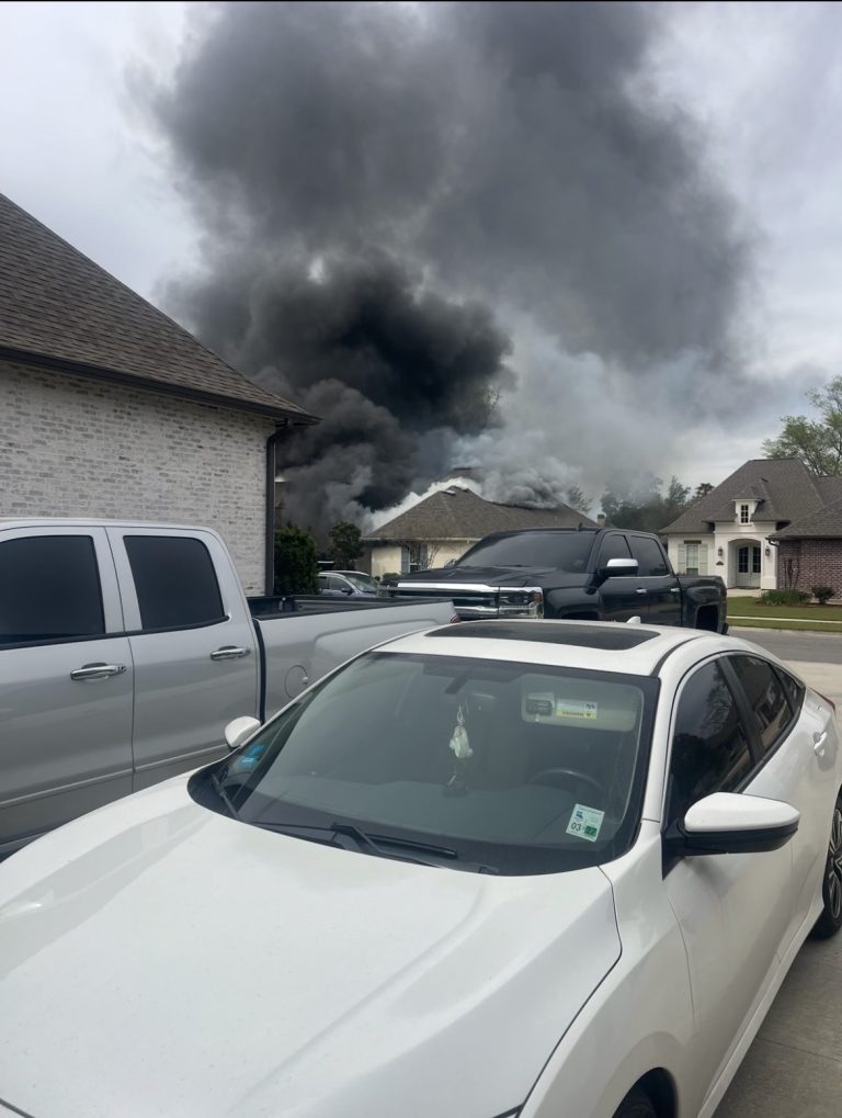 Thick black smoke rises from a house fire in a residential neighborhood. Several parked vehicles, including a white car and silver trucks, are visible in the foreground.